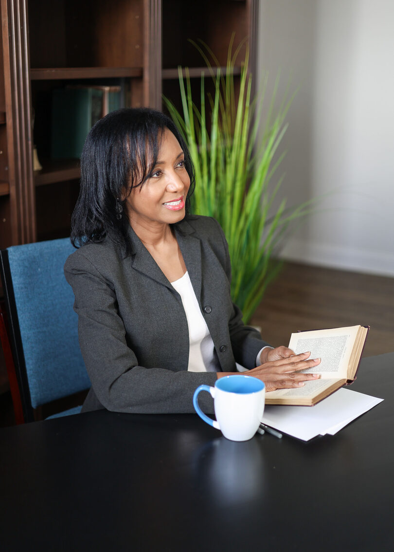 Dr. Anna-Maria Rivas McGowan seated at a table in a dark gray blazer, smiling and looking to one side with one hand on an open book beside a blue and white mug; a portrait, not a photo of her speaking